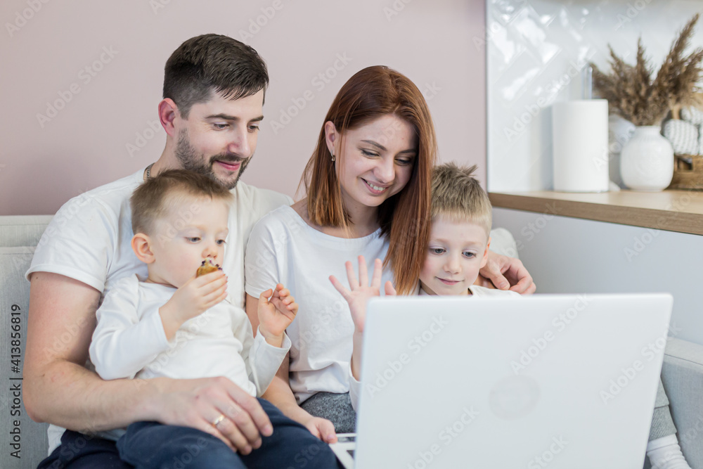 Young family, husband, wife and two young sons, sitting at home in the kitchen, and communicating via video link with family, using laptop
