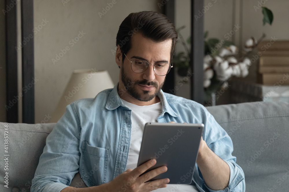Serious millennial male in glasses sit on couch focus attention on digital pad screen read electronic book online. Concentrated guy student using tablet pc for distant learning watch education video Serious millennial male in glasses sit on couch focus attention on digital pad screen read electronic book online. Concentrated guy student using tablet pc for distant learning watch education video