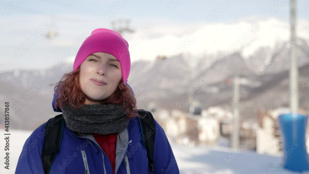 A woman in a pink hat and blue jacket stands and smiles on a city winter street against the backdrop of mountains and ski lifts. Winter resort. A woman in a pink hat and blue jacket stands and smiles on a city winter street against the backdrop of mountains and ski lifts. Winter resort.