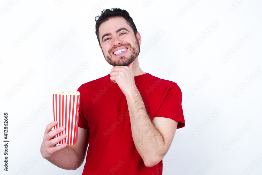 Optimistic Young handsome man in red T-shirt against white background eating popcorn keeps hands partly crossed and hand under chin, looks at camera with pleasure. Happy emotions concept. Optimistic Young handsome man in red T-shirt against white background eating popcorn keeps hands partly crossed and hand under chin, looks at camera with pleasure. Happy emotions concept.