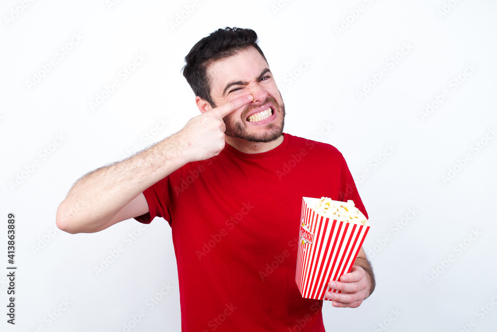 Young handsome man in red T-shirt against white background eating popcorn pointing unhappy to pimple on forehead, ugly infection of blackhead. Acne and skin problem Young handsome man in red T-shirt against white background eating popcorn pointing unhappy to pimple on forehead, ugly infection of blackhead. Acne and skin problem