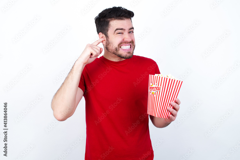 Young handsome man in red T-shirt against white background eating popcorn covering ears with fingers with annoyed expression for the noise of loud music. Deaf concept. Young handsome man in red T-shirt against white background eating popcorn covering ears with fingers with annoyed expression for the noise of loud music. Deaf concept.