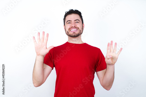 Young handsome man in red T-shirt against white background eating popcorn showing and pointing up with fingers number nine while smiling confident and happy. © Roquillo