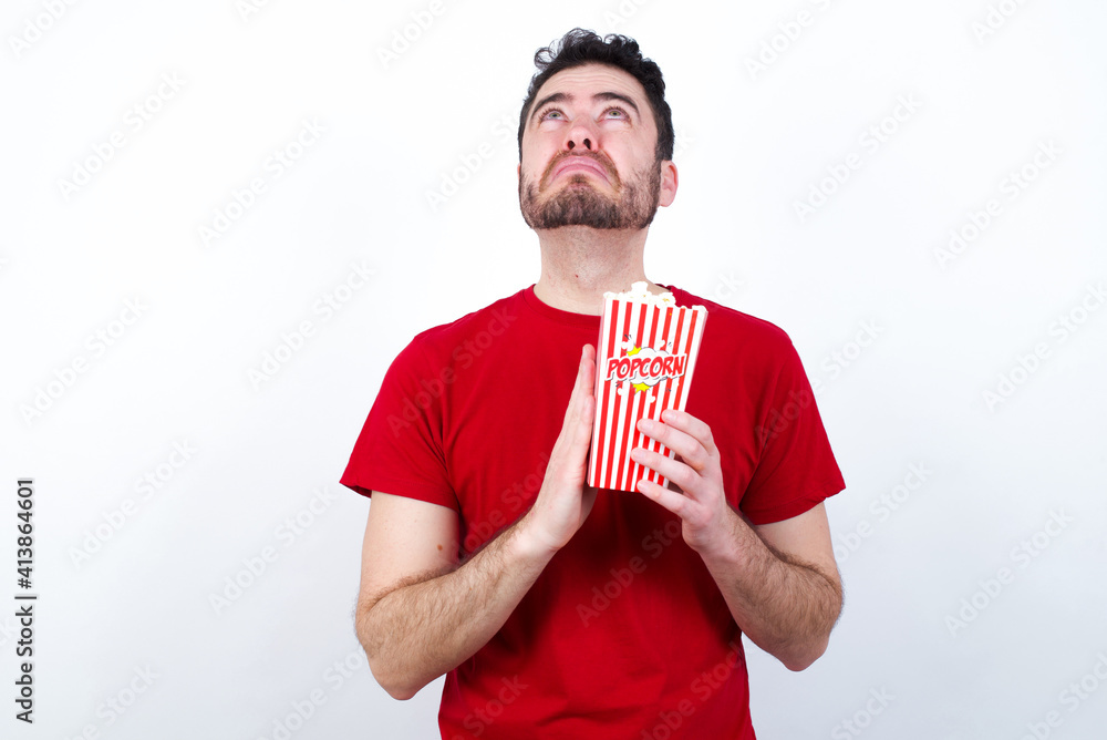 Young handsome man in red T-shirt against white background eating popcorn begging and praying with hands together with hope expression on face very emotional and worried. Please God Young handsome man in red T-shirt against white background eating popcorn begging and praying with hands together with hope expression on face very emotional and worried. Please God