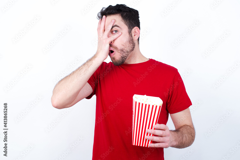 Young handsome man in red T-shirt against white background eating popcorn peeking in shock covering face and eyes with hand, looking through fingers with embarrassed expression. Young handsome man in red T-shirt against white background eating popcorn peeking in shock covering face and eyes with hand, looking through fingers with embarrassed expression.