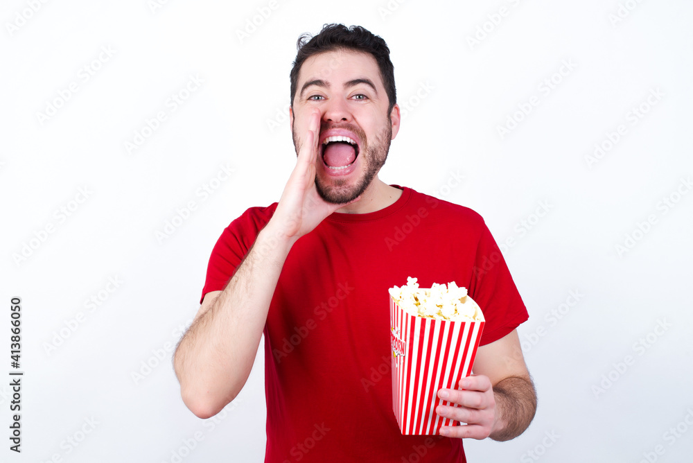 Young handsome man in red T-shirt against white background eating popcorn shouting excited to front. Young handsome man in red T-shirt against white background eating popcorn shouting excited to front.