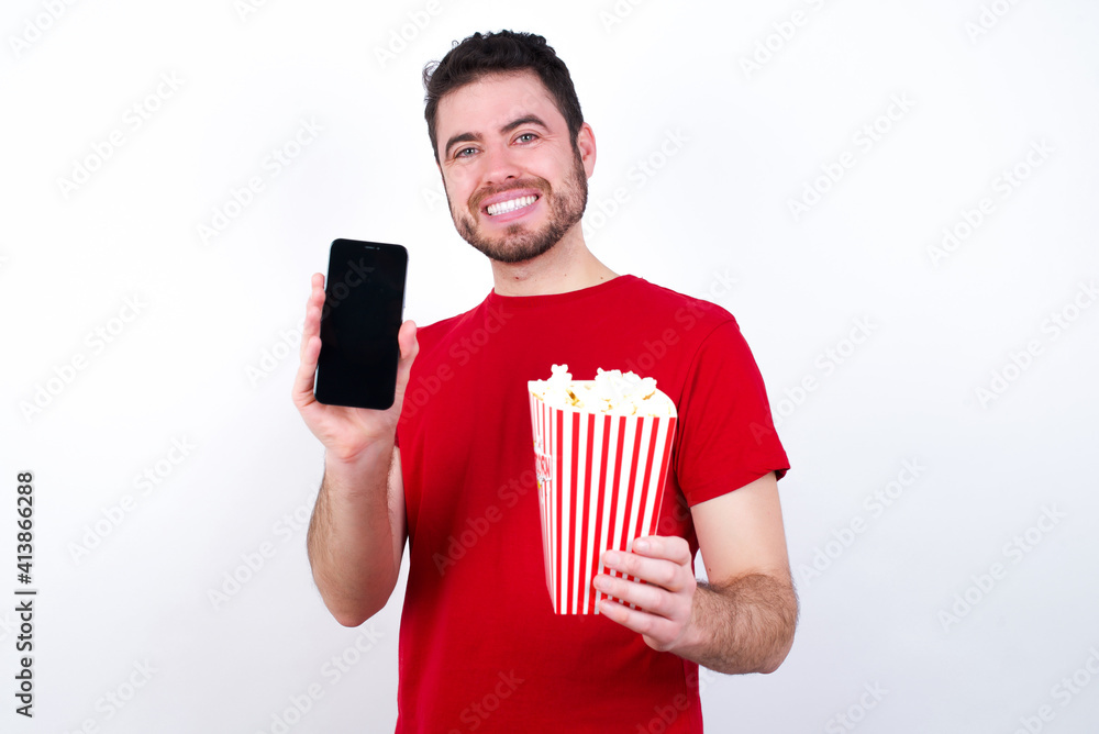 Young handsome man in red T-shirt against white background eating popcorn with a mobile. presenting smartphone. Advertisement concept. Young handsome man in red T-shirt against white background eating popcorn with a mobile. presenting smartphone. Advertisement concept.