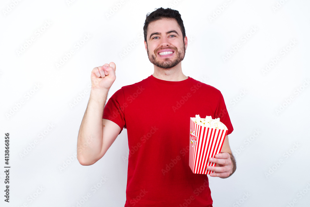 Young handsome man in red T-shirt against white background eating popcorn pointing up with fingers number ten in Chinese sign language Shi Young handsome man in red T-shirt against white background eating popcorn pointing up with fingers number ten in Chinese sign language Shi