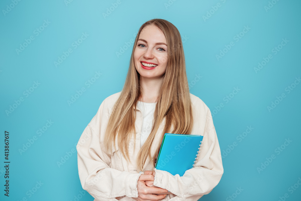 Blonde student girl holding a book, notebook smiling, looking at the camera isolated on blue background in studio Blonde student girl holding a book, notebook smiling, looking at the camera isolated on blue background in studio