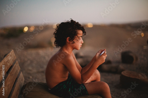 A young boy playing with his smartphone sitting on a bench in desert photo