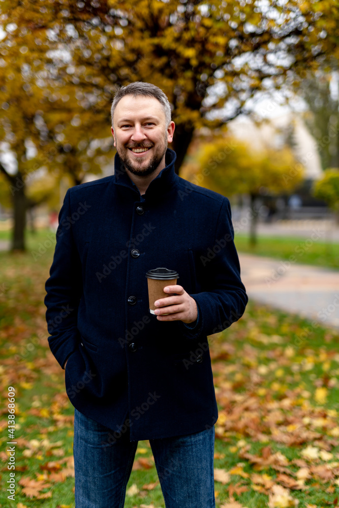 Smiling bearded male in formal clothes smiles to camera on autumn park background. Man with hot drinks in hand. Closeup. Smiling bearded male in formal clothes smiles to camera on autumn park background. Man with hot drinks in hand. Closeup.
