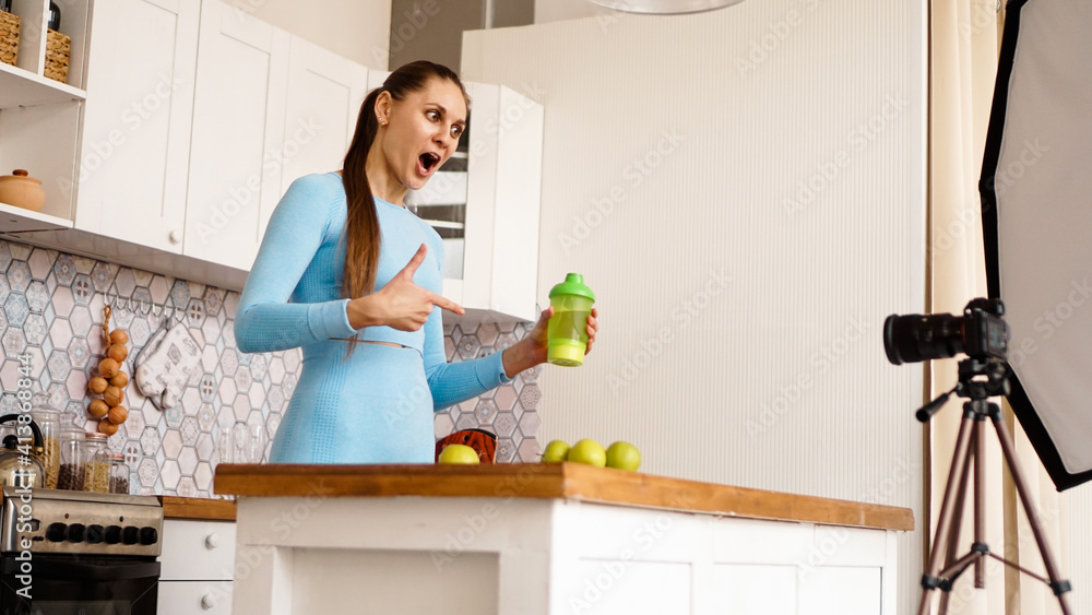 Healthy woman recording her video blog about healthy food additives while standing at kitchen. She is holding a bottle of sports nutrition and smiling Healthy woman recording her video blog about healthy food additives while standing at kitchen. She is holding a bottle of sports nutrition and smiling