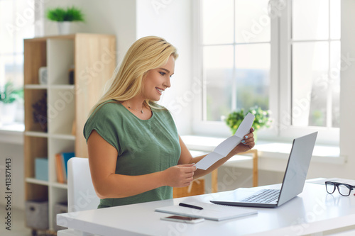 Happy young woman sitting at desk at home reading monthly statement or document notifying about business loan approval, college or university admission, winning scholarship, or getting money refund