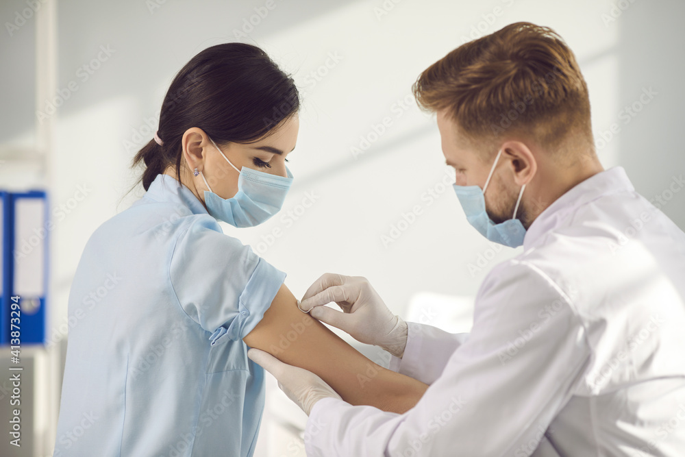 Obraz premium Man medical worker in protective face mask putting plaster to woman patient arm after coronavirus vaccination during pandemic in medical clinic, rear view. Vaccine against COVID-19 infection concept