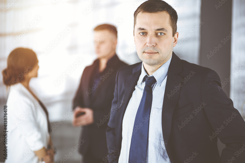 Portrait of a self-confident middle aged businessman in a blue suit, standing in a sunny modern office with his colleagues at the background. Concept of business success Portrait of a self-confident middle aged businessman in a blue suit, standing in a sunny modern office with his colleagues at the background. Concept of business success