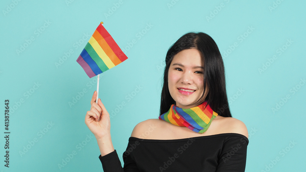 Portrait of young girl holding an LGBT flag standing against a blue green background studio. Asian LGBTQ woman with rainbow scarf on neck. look smart bright and energetic cheerful. LGBTQ concept. Portrait of young girl holding an LGBT flag standing against a blue green background studio. Asian LGBTQ woman with rainbow scarf on neck. look smart bright and energetic cheerful. LGBTQ concept.