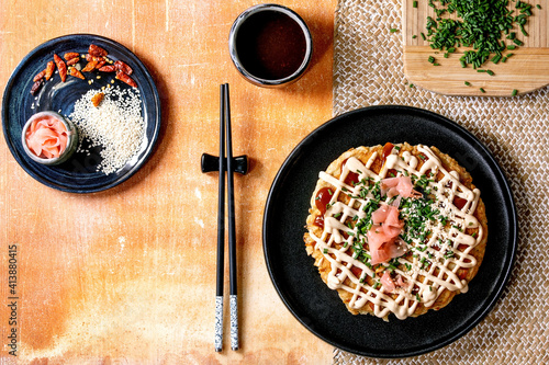 Homemade japanese fast food okonomiyaki cabbage pancake decorated by spring onion, pickled ginger, mayo sauce on black ceramic plate with chopsticks and ingredients above. Texture background. Flat lay photo