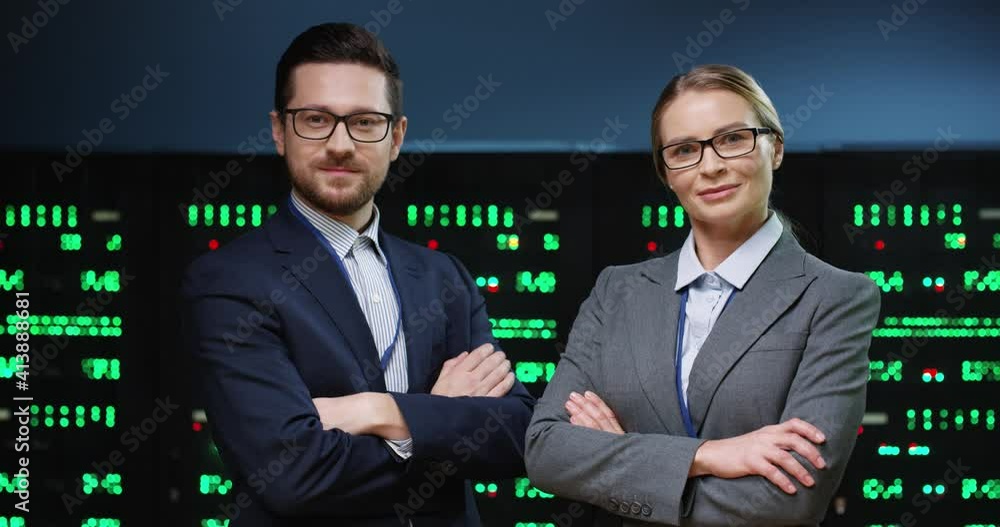 Portrait of two smart and successful programmers, man and woman, standing on background of running servers looking at camera with arms crossed. Portrait of two smart and successful programmers, man and woman, standing on background of running servers looking at camera with arms crossed.