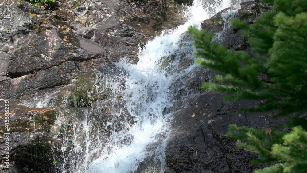 Waterfall cascades down stone wall. Sun shines on cliff and mountain stream. In foreground is spruce branch. Stones are covered with moss. Waterfall cascades down stone wall. Sun shines on cliff and mountain stream. In foreground is spruce branch. Stones are covered with moss.