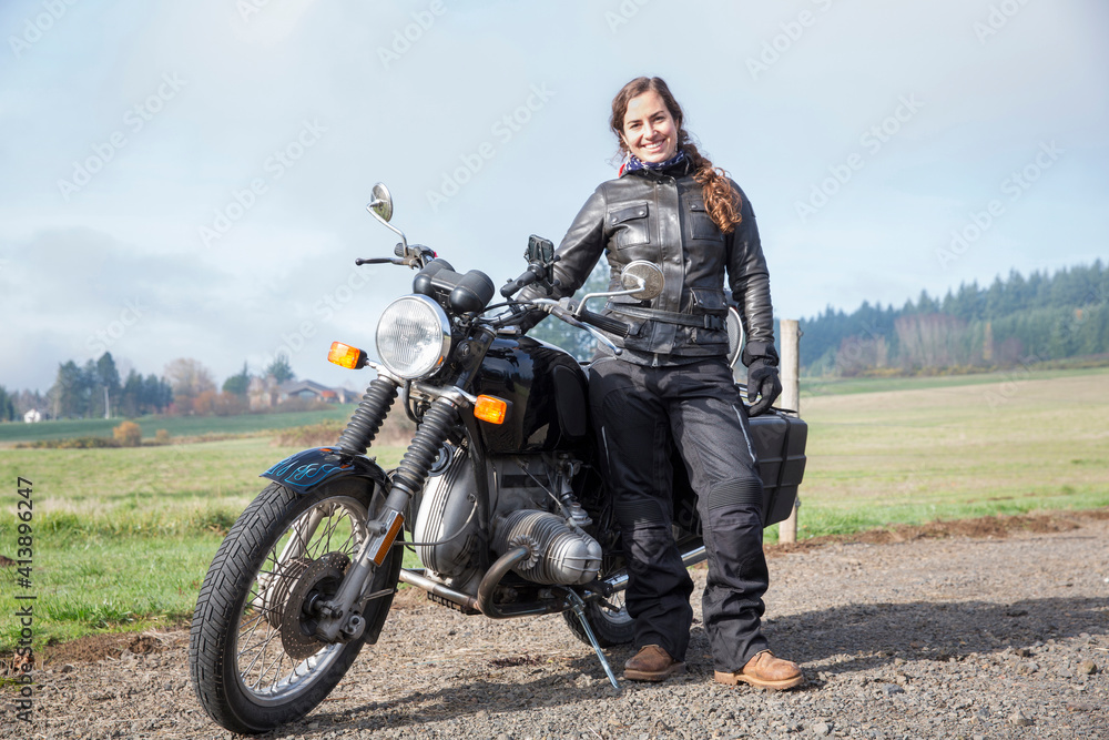 Full length portrait of female biker wearing leather jacket while standing by motorcycle on dirt road Full length portrait of female biker wearing leather jacket while standing by motorcycle on dirt road