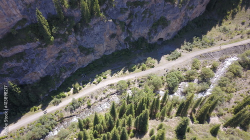 Top view of the cave, forest, rocks and road. A group of tourists walking along the trail. There is a huge cave in the rock. Green coniferous forest, trees and grass grow on the rocks. The river runs.