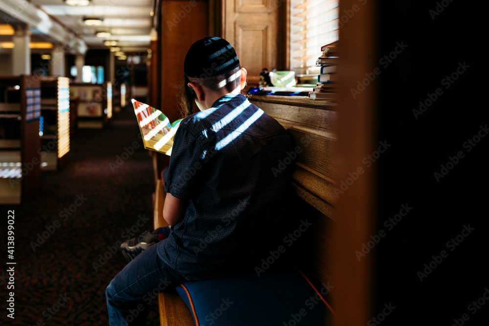 Rear view of boy reading book by window in library Rear view of boy reading book by window in library
