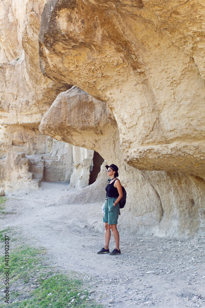 woman traveler with a backpack and a hat stands next to a rock in the Crimea in the summer