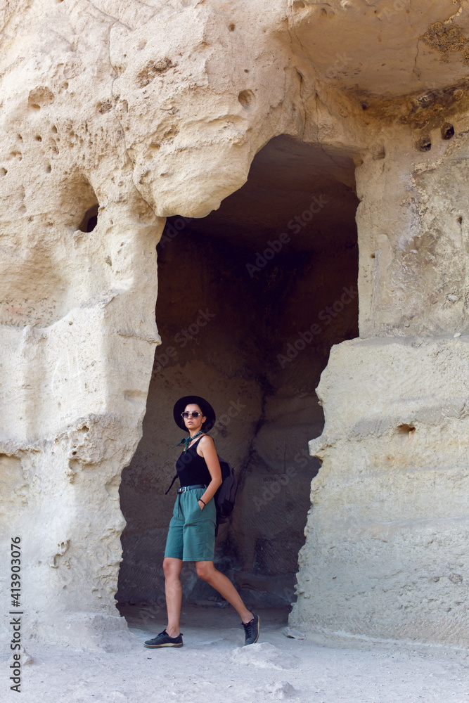 woman traveler with a backpack and a hat stands next to a rock in the Crimea in the summer