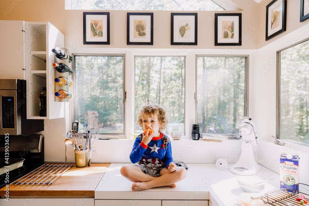 Full length of cute boy eating fruit while sitting on kitchen counter against windows at home Full length of cute boy eating fruit while sitting on kitchen counter against windows at home