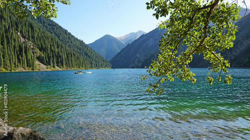 Fototapeta Naklejka Na Ścianę i Meble -  Mountain lake Kolsay with floating boats. View of green hills, fir trees, grass and water. Boats with tourists float on the lake. People are resting. The water of a mountain lake is green.