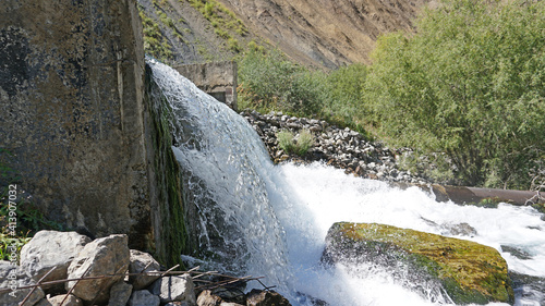 Draining the water pressure into the river. The water flows like a waterfall and flows into the river. There are concrete walls around the edges  and iron pipes are visible. Trees grow in the distance