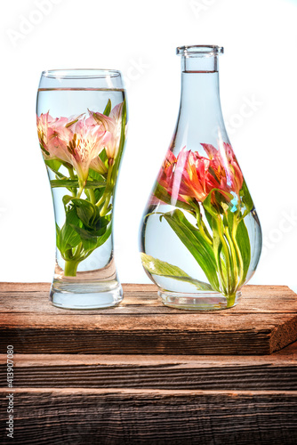 Cut flowers and pink lilies in a vase filled with water on a barn wood table in front of a white background