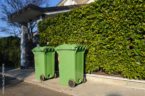 Bidoni carrellati verdi all’esterno di un’abitazione davanti a una siepe photo