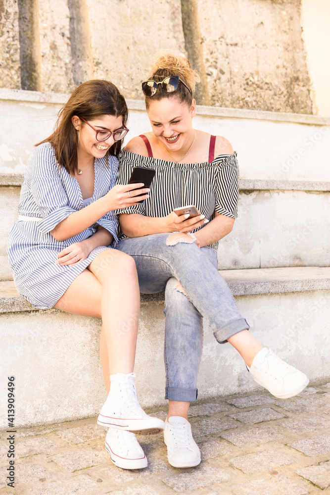 Full length of smiling female tourists using phone while sitting on steps Full length of smiling female tourists using phone while sitting on steps