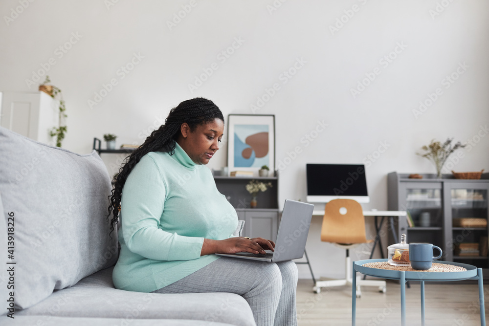 Side view portrait of curvy African American woman using laptop while browsing internet sitting on couch in minimal home interior, copy space Side view portrait of curvy African American woman using laptop while browsing internet sitting on couch in minimal home interior, copy space