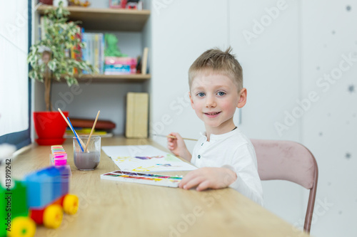 Little blond boy draws in his room sitting at a table, having fun
