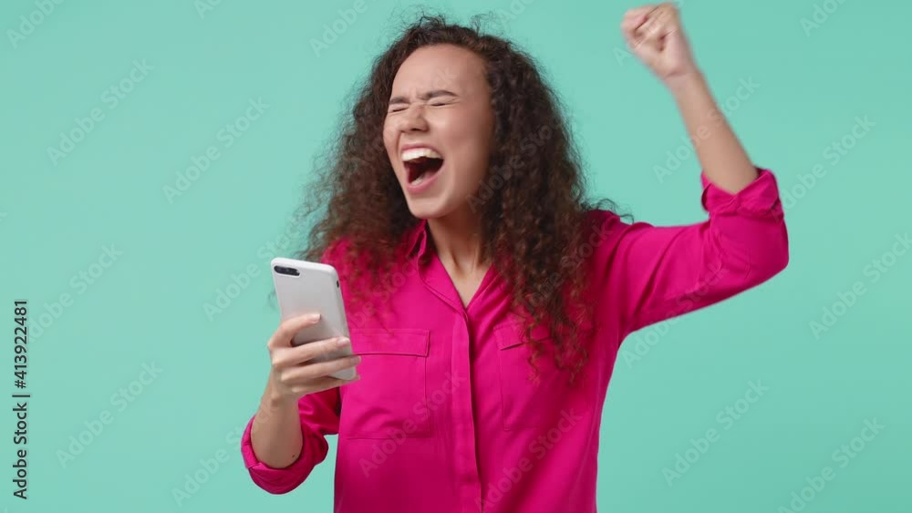 Surprised joyful young african american woman 20s in pink shirt isolated on blue turquoise background studio. People lifestyle concept. Using mobile cell phone typing browsing doing winner gesture Surprised joyful young african american woman 20s in pink shirt isolated on blue turquoise background studio. People lifestyle concept. Using mobile cell phone typing browsing doing winner gesture