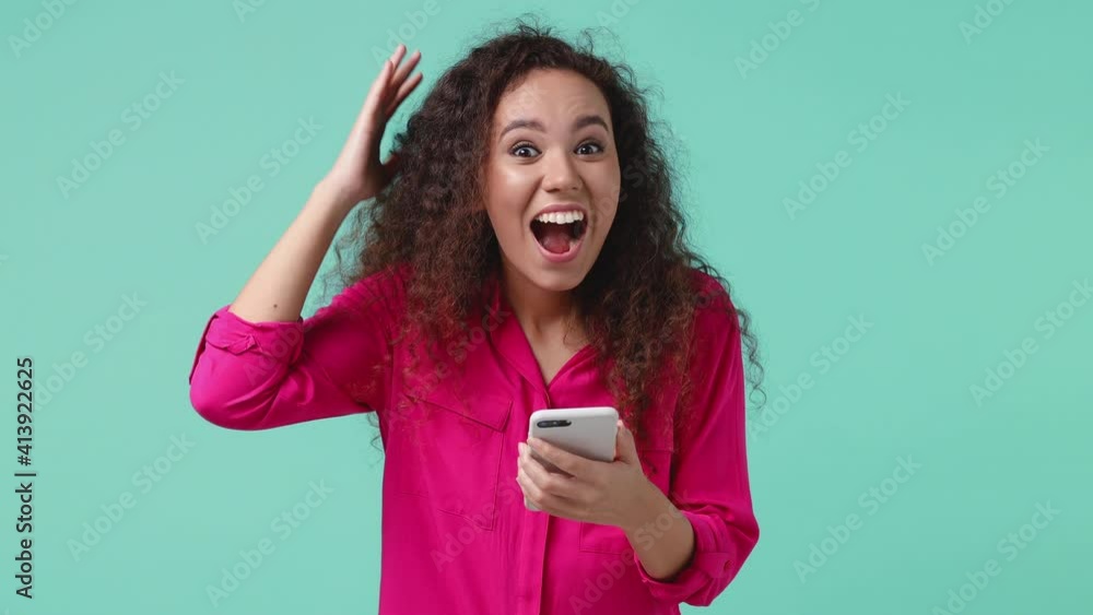 Surprised amazed young african american woman 20s in pink shirt isolated on blue turquoise background studio. People lifestyle concept. Pointing index finger on mobile cell phone doing winner gesture Surprised amazed young african american woman 20s in pink shirt isolated on blue turquoise background studio. People lifestyle concept. Pointing index finger on mobile cell phone doing winner gesture