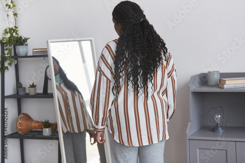 Minimal back view portrait of overweight African American woman looking in mirror while trying on clothes at home, copy space