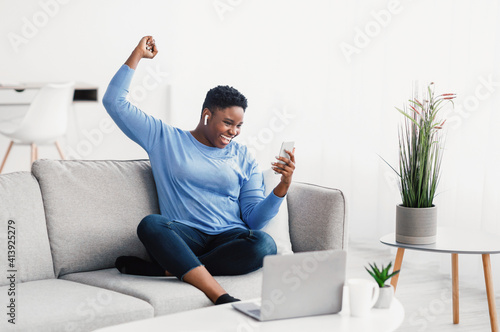 Excited black lady wearing headphones using phone © Prostock-studio