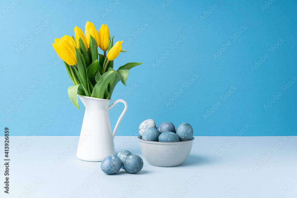 Bouquet of yellow fresh tulips in white jug, gray plate with painted Easter eggs on blue background. Bouquet of yellow fresh tulips in white jug, gray plate with painted Easter eggs on blue background.
