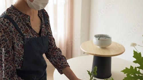 Mujer con mascarilla tocando plantas. Mesa con jarra blanca