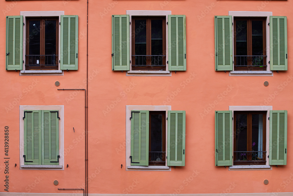 Pink building faced with six green windows, one of which is closed, Italy, Alps