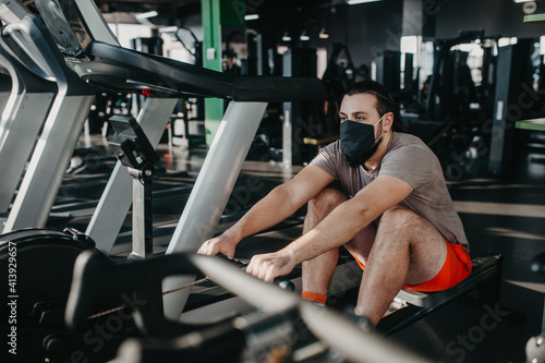 An active man with beard in a protective medical mask training in gym using rowing machine. Cardio