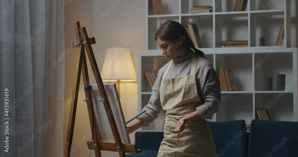 young woman is painting at home at evening, using professional easel, canvas, paint and brush in apartment young woman is painting at home at evening, using professional easel, canvas, paint and brush in apartment