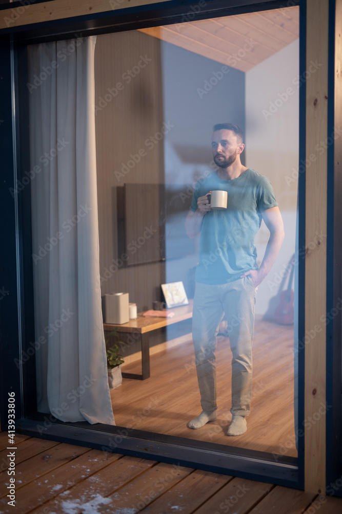 Young bearded man holding mug with hot drink and looking through large window