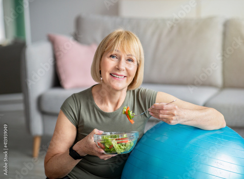 Exercise and healthy diet concept. Senior woman with fitball eating fresh vegetable salad at home