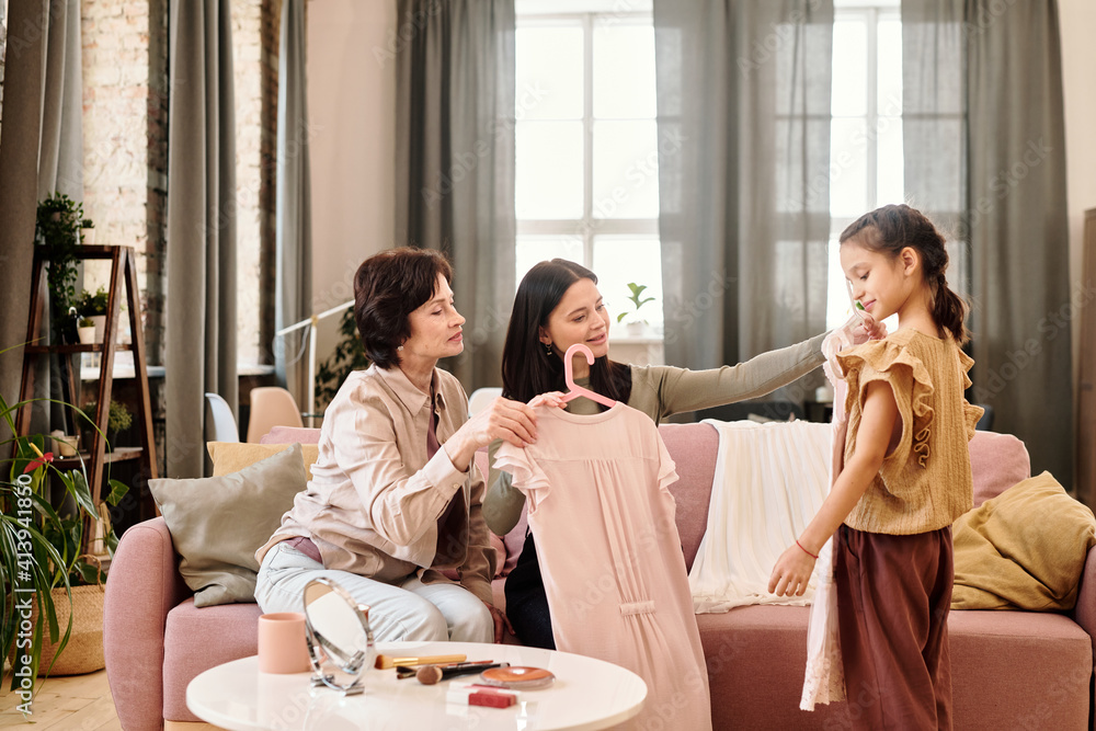 Two women sitting on couch in living-room and showing new dresses to little girl