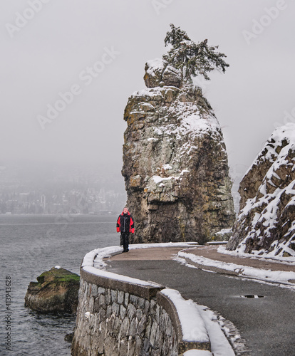 Man walking on seawall by Siwash Rock in Stanley Park in winter snowy foggy weather. Vancouver. British Columbia. Canada  photo