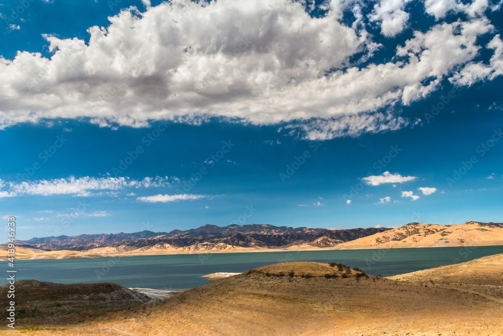calm lake under a blue sky with cotton like clouds above the sky and surrounded by desert like terrain in Central Valley California.